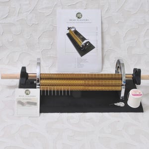May include: A Read Pleaters fabric pleating machine featuring gold-colored rollers on a black base, accompanied by an instruction booklet, a small card with the Read Pleaters logo, a spool of white thread, and two small metal needles.