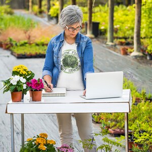 May include: A woman in a blue denim shirt and white t-shirt with the text "Grow Food Not Lawns" is working on a laptop at a table in a greenhouse. There are potted plants on the table and in the background.