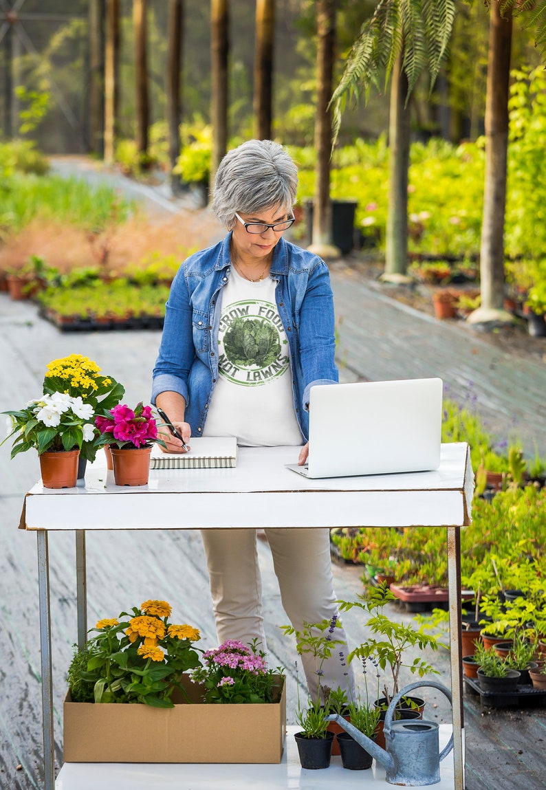 May include: A woman in a blue denim shirt and white t-shirt with the text "Grow Food Not Lawns" is working on a laptop at a table in a greenhouse. There are potted plants on the table and in the background.