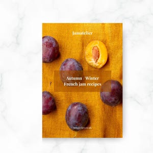 May include: A cookbook cover with the text "Autumn / Winter French jam recipes" and the website address "jamatelier.co.uk". The cover is on a yellow cloth and features several purple plums, one cut open to reveal the yellow flesh.