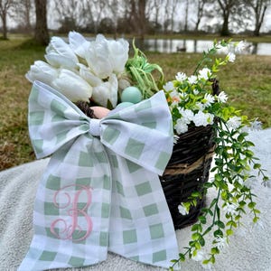 Könnte beinhalten: Ein geflochtener Korb gefüllt mit weißen Tulpen, künstlichen Eiern und Grünzeug. Eine große, hellgrüne und weiße Vichy-Schleife mit einem rosa gestickten "B" ist an dem Korb befestigt. Der Korb steht draußen auf einer weißen Decke.