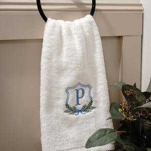 May include: White hand towel with a blue embroidered initial "P" inside a shield design, surrounded by a wreath and bow. The towel hangs from a black metal ring in a bathroom setting.