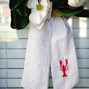 May include: A magnolia wreath featuring large white flowers and dark green leaves. A striped fabric bow with a red embroidered lobster is attached. The background is a light blue shutter.