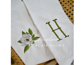 May include: Two white fabric towels featuring embroidery. One towel displays a green letter H, while the other features a detailed white magnolia flower with green leaves. The text Handmade BY BLAIR PALMA is overlaid in white across the center.