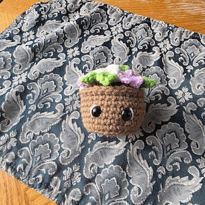 Peut inclure: Un petit pot de plante crocheté marron avec deux grands yeux de sécurité noirs et brillants. Le pot contient un feuillage crocheté dans des tons de vert, blanc et violet clair, imitant une petite plante en pot à l'aspect sympathique.