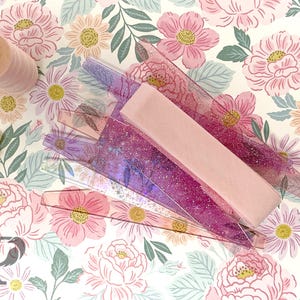 May include: A collection of sewing supplies, including pink thread spools, a light pink ribbon, and several clear, glitter-infused rulers. The items are arranged on a floral patterned fabric with pink and white flowers.