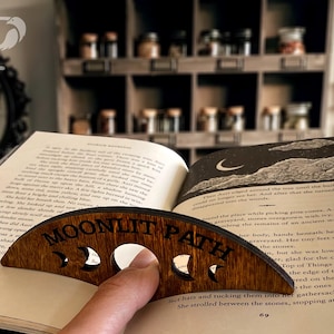 May include: A wooden book holder with the words "MOONLIT PATH" engraved on it. The holder is shaped like a crescent moon and has cutouts of different moon phases.