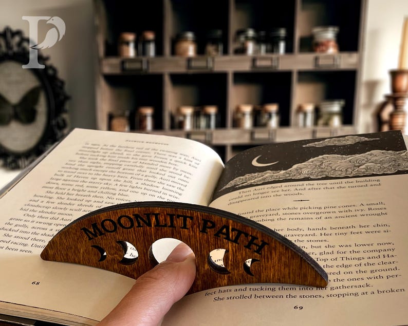 May include: A wooden book holder with the words "MOONLIT PATH" engraved on it. The holder is shaped like a crescent moon and has cutouts of different moon phases.