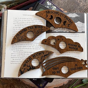 May include: Set of six wooden bookmarks with different designs and sayings. The designs include a crescent moon, a cloud, a rainbow, a mountain, and a star. The sayings include "Moonlit Path", "Reading is Magical", "Wild Hunt", "Chasing the Wind", "Time to Be", and "A Unicorn".