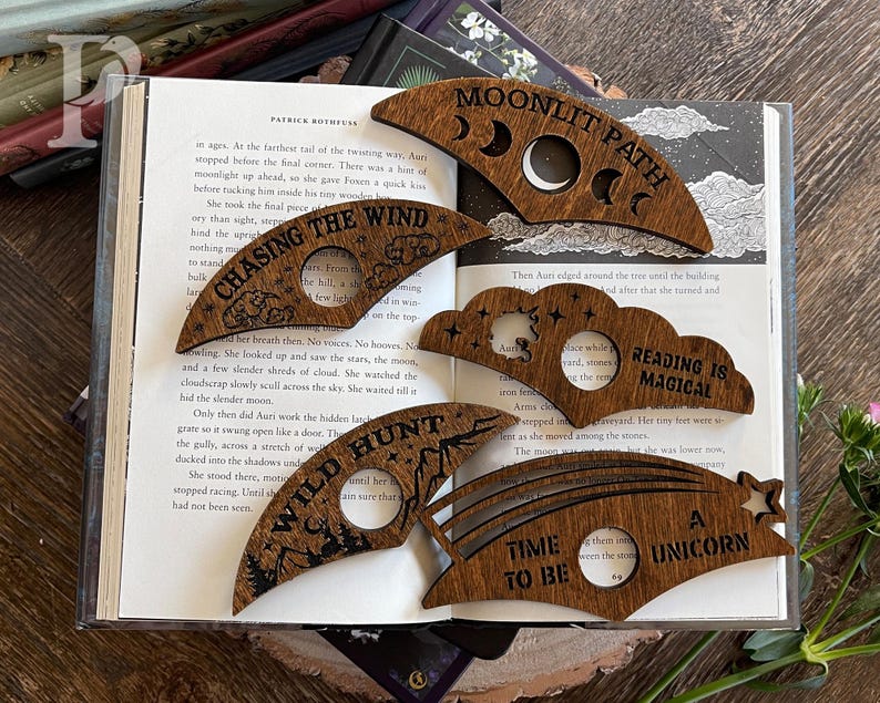 May include: Set of six wooden bookmarks with different designs and sayings. The designs include a crescent moon, a cloud, a rainbow, a mountain, and a star. The sayings include "Moonlit Path", "Reading is Magical", "Wild Hunt", "Chasing the Wind", "Time to Be", and "A Unicorn".