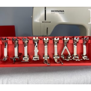 May include: A red plastic organizer holding various sewing machine presser feet. The feet are silver and white, with different shapes and functions. A Bernina sewing machine is in the background.