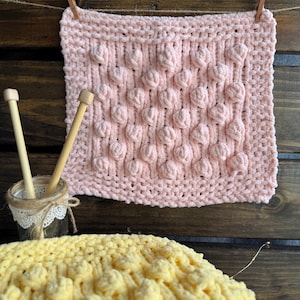 Puede incluir: Una manta tejida a mano, cuadrada, rosa, con un patrón de burbujas en relieve texturizado, colgada de un tendedero con una pinza de madera. Dos agujas de tejer descansan en un pequeño tarro de cristal. Una manta tejida amarilla está en primer plano.
