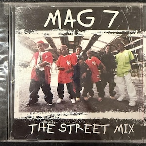 Puede incluir: Caja de CD con el título "MAG 7" en blanco, con fuente manuscrita. La portada presenta un grupo de jóvenes con ropa roja y negra. El texto "THE STREET MIX" está en la parte inferior.