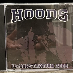 Op de afbeelding: Een doorzichtig plastic CD-doosje met een muziekalbum. De hoes toont het woord HOODS in grote witte letters met een donkere omlijning bovenaan en de tekst DEMONSTRATION 2005 in het wit onderaan over een donkere grafische afbeelding.