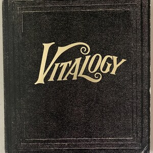 Puede incluir: Un libro negro con la palabra "VITALOGY" en letras doradas. El libro tiene una cubierta texturizada y un borde decorativo. El título está en una fuente estilizada.