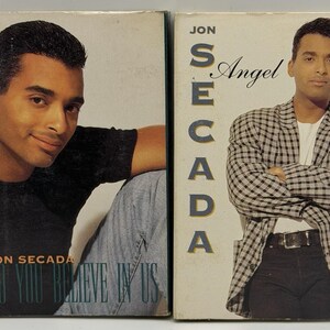 May include: Two vintage music album covers featuring singer Jon Secada. One cover shows Secada in a black shirt and jeans, with the text "Do You Believe In Us." The other cover displays Secada in a checkered jacket, with the text "Angel."