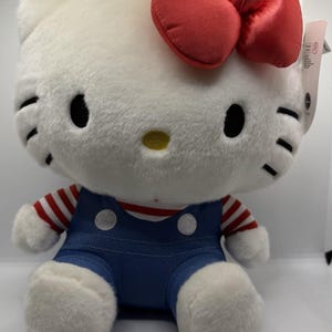 May include: A plush Hello Kitty doll with a white body, black eyes, and a yellow nose. She wears a red bow and blue denim overalls with white buttons. Her sleeves are striped red and white.