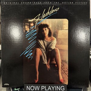 May include: A vinyl record album cover for the movie "Flashdance." The cover is black with the movie title in yellow script and a woman in a gray sweater and red heels. Blue diagonal lines are on the left side of the title.