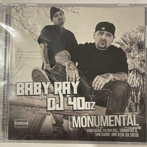 May include: A CD case featuring two men in black clothing. The album title "Monumental" is displayed with the artists' names "Baby Ray and DJ 40oz." The cover includes a parental advisory warning and lists featured artists.