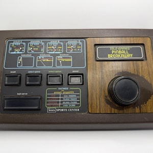 Puede incluir: Una consola vintage Sears Sports Center Tele-Games Pinball Breakaway. La consola marrón presenta un panel de vetas de madera, un gran dial negro y varios botones. La pantalla muestra gráficos del juego e información de puntuación. El texto "PINBALL BREAKAWAY" es visible.