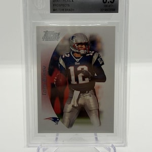 Könnte beinhalten: Eine Tom Brady Footballkarte von 2005 Topps Draft Picks & Prospects, bewertet mit 8,5, in einem Schutzhalter. Die Karte zeigt Tom Brady in seinem New England Patriots-Trikot, mit der Nummer 12 sichtbar.