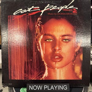 May include: The album cover for the "Cat People" original soundtrack. The cover features a woman's face with green eyes and a red and orange color scheme. The text "Cat People" and "Music by Giorgio Moroder" are visible.