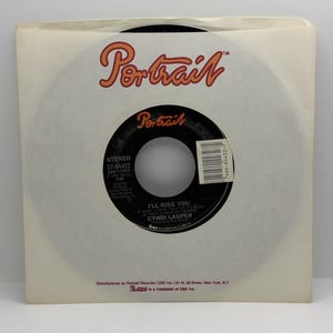 Puede incluir: Un disco de vinilo de 45 RPM vintage de la canción "I'll Kiss You" de Cyndi Lauper en una funda de papel blanca. La etiqueta del disco es negra con texto blanco y el logotipo "Portrait". La funda tiene el logotipo "Portrait" en naranja.