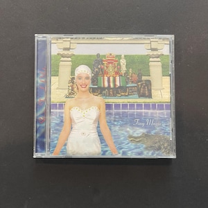 Puede incluir: Una caja de CD con una mujer con un vestido blanco y un tocado en una piscina. El fondo muestra una exhibición decorativa y un cocodrilo. El texto "Tiny Music" es visible en la parte inferior derecha.