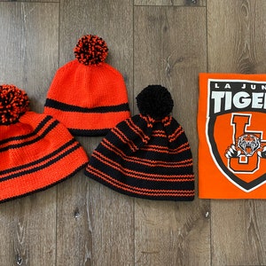 Op de afbeelding: Drie zwarte en oranje gestreepte gebreide mutsen met pompoenen en een La Junta Tigers T-shirt.