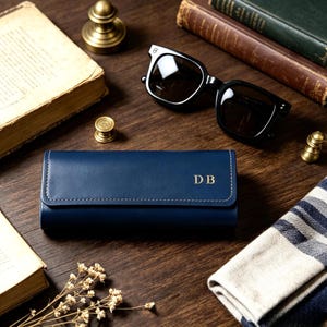 May include: A rectangular navy blue leather glasses case featuring a flap closure with white stitching and the gold embossed initials DB on the front. The case has a smooth finish and is positioned horizontally on a dark wooden surface.
