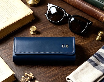May include: A rectangular navy blue leather glasses case featuring a flap closure with white stitching and the gold embossed initials DB on the front. The case has a smooth finish and is positioned horizontally on a dark wooden surface.