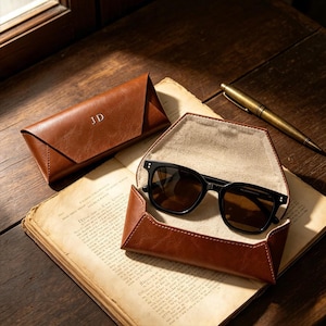 Könnte beinhalten: Eine Sonnenbrille mit schwarzem Rahmen und dunklen Gläsern in einem offenen, dreieckigen, braunen Etui in Lederoptik. Ein passendes geschlossenes Etui mit den eingeprägten Buchstaben JD und ein messingfarbener Stift liegen daneben.