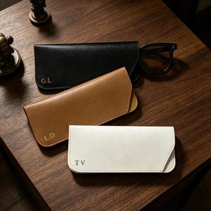 Peut inclure: Trois étuis à lunettes rectangulaires en noir, beige et blanc. L'étui noir porte les initiales dorées GL, l'étui beige les initiales dorées LD et l'étui blanc les initiales vertes TV. Une paire de lunettes noires est visible dans l'étui noir.