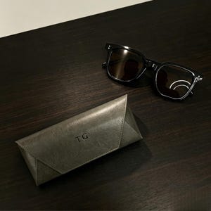 Könnte beinhalten: Eine Sonnenbrille mit schwarzem Rahmen und dunklen Gläsern neben einem dreieckigen, grauen, strukturierten Brillenetui. Das Etui hat einen spitz zulaufenden Klappverschluss und die Buchstaben TG sind in einer serifenlosen Schrift eingeprägt.