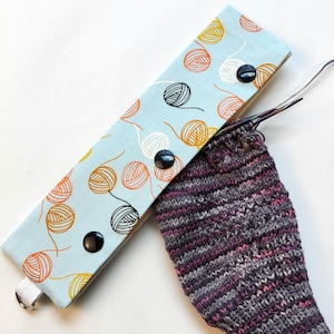May include: A light blue knitting needle case with a pattern of yarn balls in orange, yellow, and black. The case is closed with three black buttons. A partially knitted sock in shades of purple, gray, and pink is next to the case.