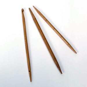Könnte beinhalten: Drei Häkel- und Strickwerkzeuge aus Holz liegen auf einer weißen Oberfläche. Links befindet sich ein Häkelhaken mit gebogenem Ende. Rechts sind zwei Stricknadeln mit spitzen Enden. Die Werkzeuge sind aus Holz.