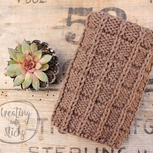 May include: A brown knitted phone case with a cable knit pattern. The case is lying on a wooden surface with a small succulent plant in a pot.