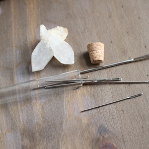May include: A glass tube filled with several silver sewing needles, a cork stopper, and a clear quartz crystal on a wooden surface.