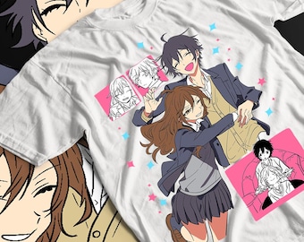 May include: White t-shirt featuring a colorful anime-style illustration of two characters embracing, with additional character portraits and star accents. The design includes a male and female character in school uniforms.