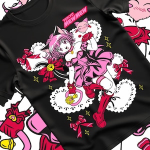 Może przedstawiać: Czarna koszulka z różowowłosą postacią z anime w różowej sukience i butach, z kocimi uszami i ogonem. Projekt zawiera napis "TOKYO MEWMEW" oraz elementy dekoracyjne, takie jak kokardy, gwiazdy i serca.