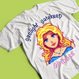 May include: White t-shirt featuring a pixelated illustration of a woman with blonde hair and blue eyes. The shirt has the words "gaslight gatekeep" in blue and "girlboss" in pink.