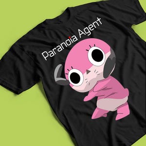 May include: Black t-shirt with a pink cartoon character with large black eyes and a grey ear. The white text "Paranoia Agent" is printed above the character. The shirt is made of a soft material.