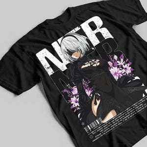 Peut inclure: T-shirt noir avec un graphisme d'un personnage aux cheveux blancs et un bandeau sur les yeux, avec les lettres "N" "I" "E" "R" en blanc. Le personnage porte une robe noire et est entouré de fleurs violettes. Texte en bas du t-shirt.