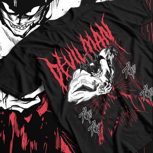 May include: Black t-shirt featuring a graphic of a muscular, horned figure with the text "DEVILMAN" in red. The design includes red splatter effects and Japanese sound effects. The shirt has a crew neck and short sleeves.
