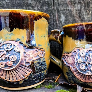 May include: Two ceramic mugs with a steampunk design. The mugs are brown, yellow, and blue with a copper-colored gear design.