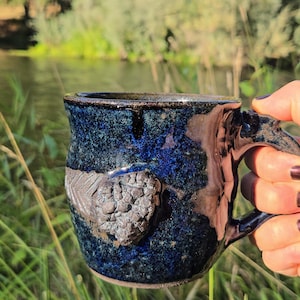 May include: A blue ceramic mug with a textured design, held by a hand with dark nail polish. The mug is in focus, while the background is blurred and shows a river and green foliage.
