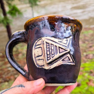 May include: A dark brown ceramic mug with a white geometric design. The mug has a rounded body and a handle.
