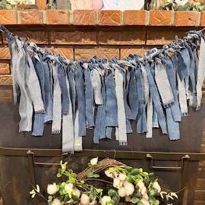 May include: A denim garland with alternating strips of light and dark blue fabric hangs in front of a brick fireplace. The garland is suspended from a thin, natural-colored string. Below the garland is a black metal fireplace screen.