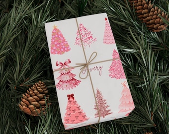 May include: A wrapped gift with a white background and pink Christmas tree patterns. The gift is tied with twine and features the name "Avery" in cursive. The gift is surrounded by pine needles and pine cones.