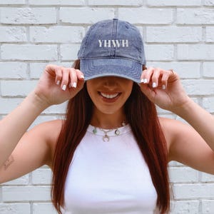 May include: A denim blue baseball cap with the white letters YHWH across the front. The hat is worn by a person in front of a white brick wall. The person is wearing a white tank top and a necklace.